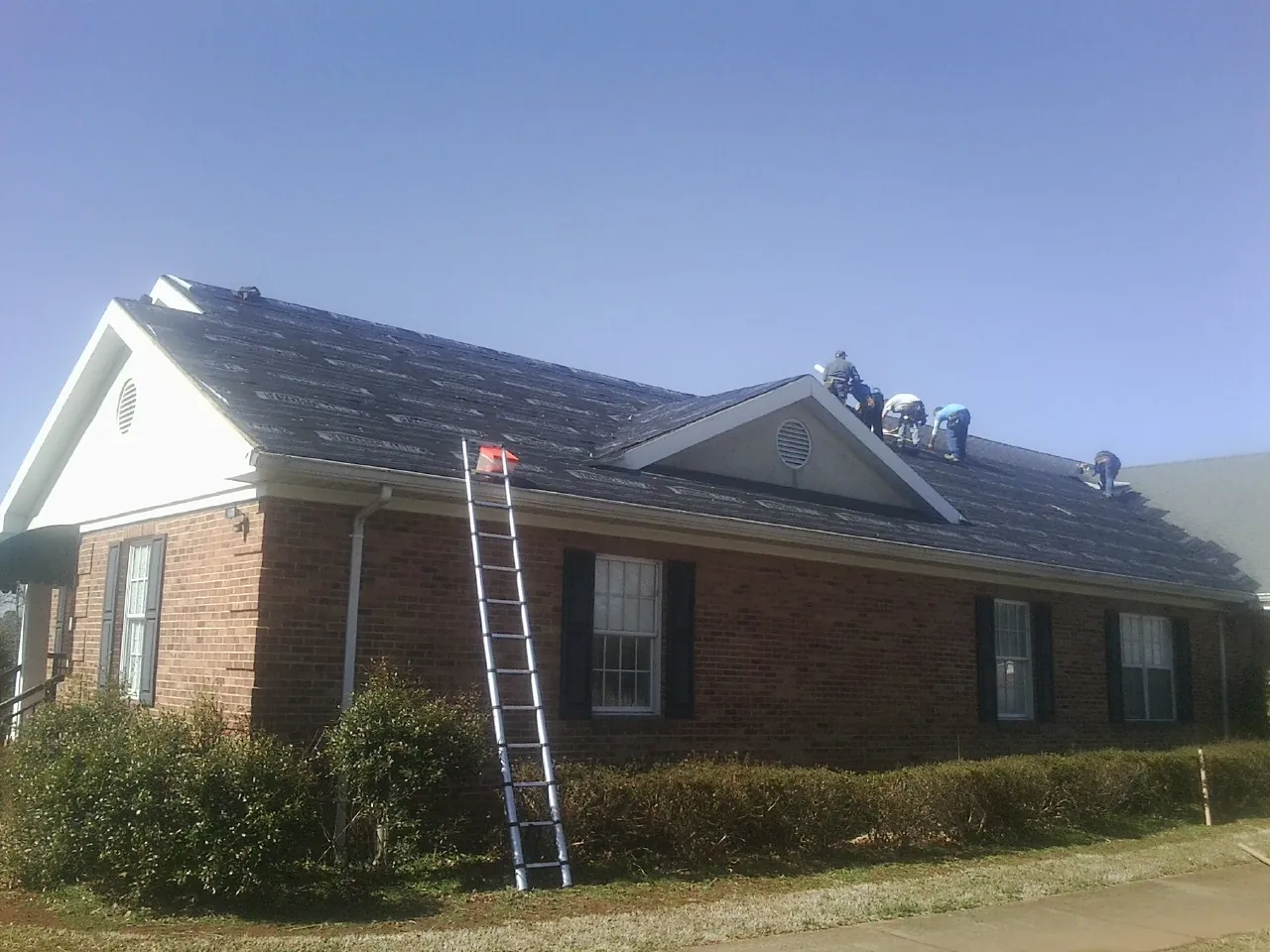 Crew tearing shingles off a brick ranch roof with a ladder set — Emergency Roof Repair work by our crew in West Leyden