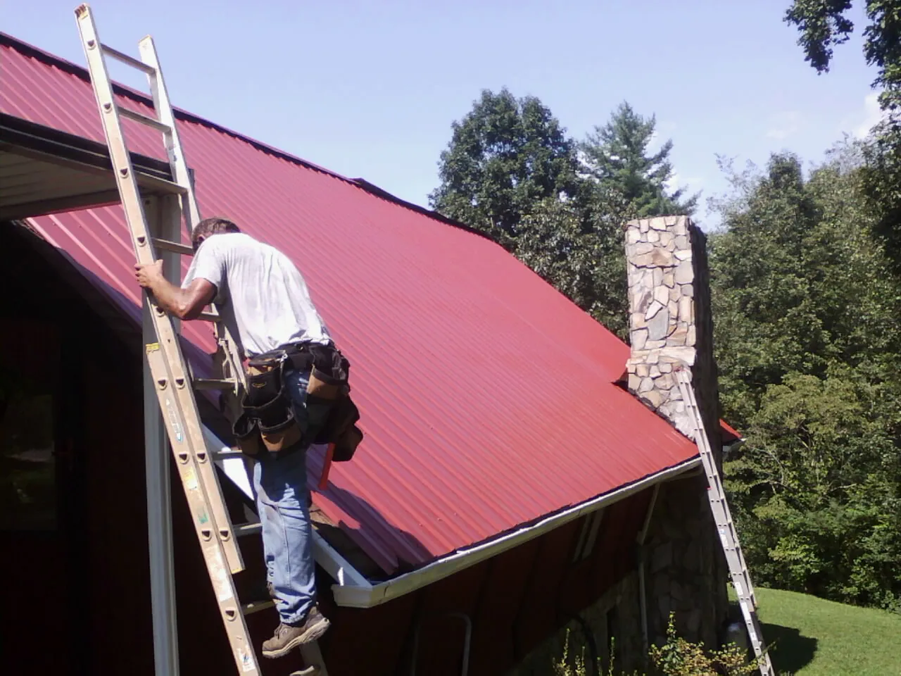 Roofer on a ladder beside a red metal roof and stone chimney — Soffit & Fascia Repair by Roofing Brothers in West Leyden